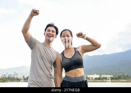 A cheerful, active Asian couple smiles at the camera, raises their fists in triumph, celebrating their success after reaching their running target fol Stock Photo