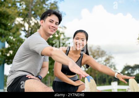 An active, sporty Asian couple in sportswear smiles at the camera while stretching their legs and warming up before running, enjoying a morning workou Stock Photo