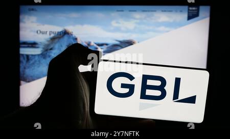 Person holding mobile phone with logo of company Groupe Bruxelles Lambert S.A. (GBL) on screen in front of web page. Focus on phone display. Stock Photo