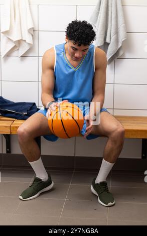 Sitting on bench, basketball player in uniform holding ball in locker room Stock Photo