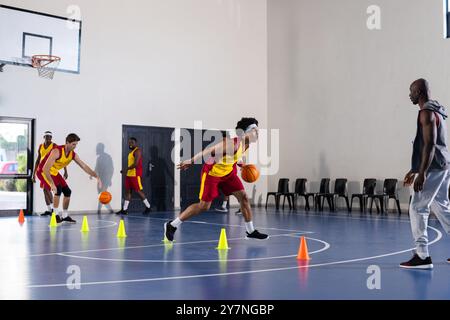 Practicing basketball drills, players dribbling around cones in gym with coach Stock Photo