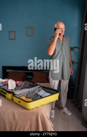 Senior man standing near open suitcase with clothes, speaking on smartphone, booking travel tour on phone from home. Copy space Stock Photo