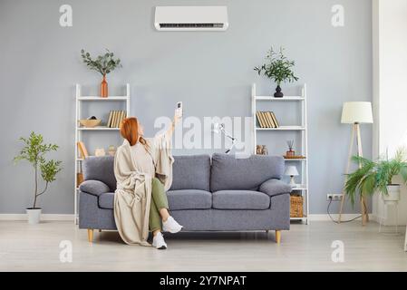 Girl wrapped in blanket switching on, off air conditioner, blowing warm air room temperature control Stock Photo