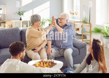 Happy senior old parents giving high five playing chess with adult children at home. Stock Photo