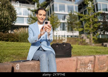 Businesswoman is using a smartphone while working remotely, taking advantage of the warm weather and sunshine to work outdoors in an urban park Stock Photo