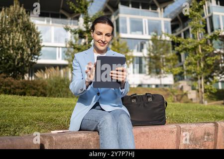 Businesswoman is using her digital tablet to work remotely while sitting outside on a bench enjoying a sunny day Stock Photo