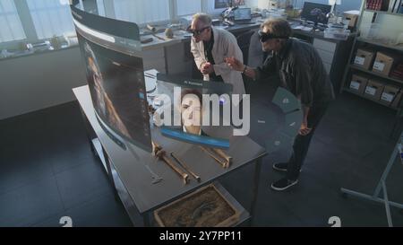 Modern archaeological lab: Two archaeologists in VR headsets study fossil skeleton using augmented reality technology. VFX graphics of virtual display with AI assistant and 3D modeling app interface. Stock Photo