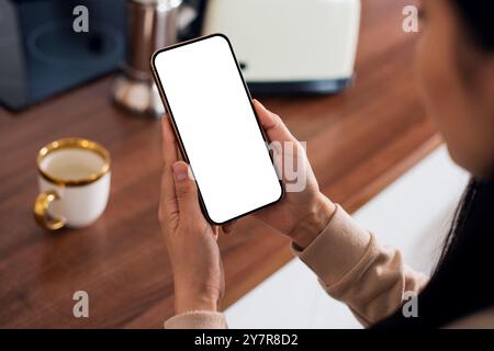 UnrecognizableAsian woman stands in the kitchen and using a smartphone mockup with a white blank screen Hands at work with Digital Technology Pov View Stock Photo