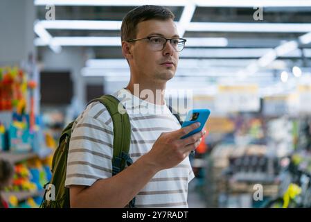 Man in striped shirt with backpack uses his smartphone while shopping in brightly lit store, appearing focused. Stock Photo
