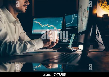 Male trader holding coffee cup while analyzing stock market data on multiple computers at night office Stock Photo