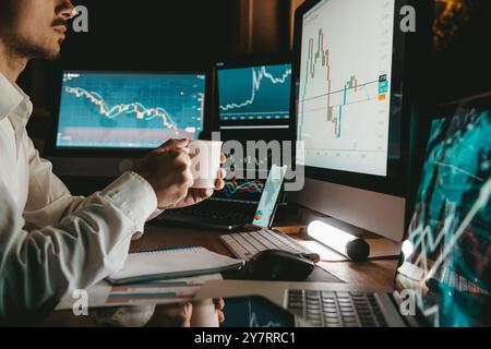 Male trader holding coffee cup while analyzing stock market data on multiple computers at night office Stock Photo