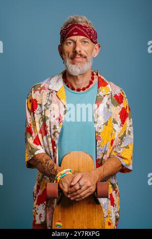 Hipster mature man in stylish shirt leaning on longboard and looking at camera against blue background Stock Photo