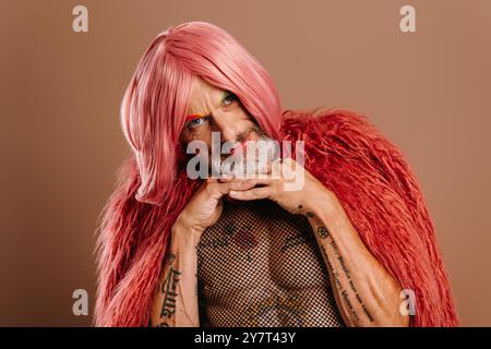 Bearded mature gay man in pink wig leaning face on hands against brown background Stock Photo
