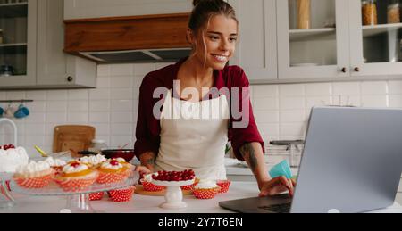 Beautiful female baker using laptop while making cakes and muffins on the kitchen Stock Photo