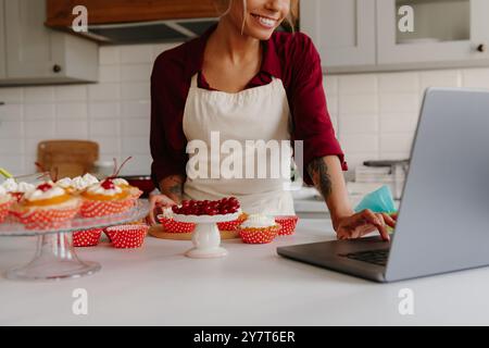 Confident female baker using laptop while making cakes and muffins on the kitchen Stock Photo