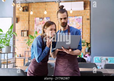 Young man and woman workers owners in aprons using laptop, inside restaurant cafe Stock Photo