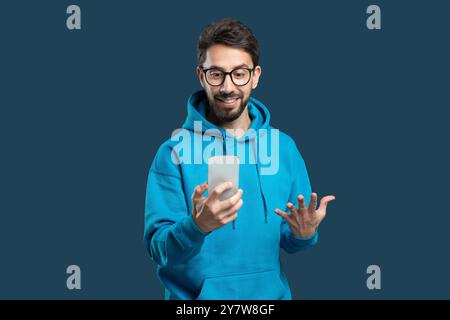 Man in a blue hoodie gestures excitedly while using a smartphone indoors Stock Photo