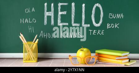 Blackboard with word HELLO in different languages and stationery on table in classroom Stock Photo