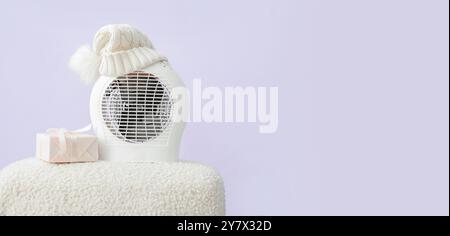Electric fan heater, hat and gift box on pouf against lilac background with space for text. Concept of heating season Stock Photo
