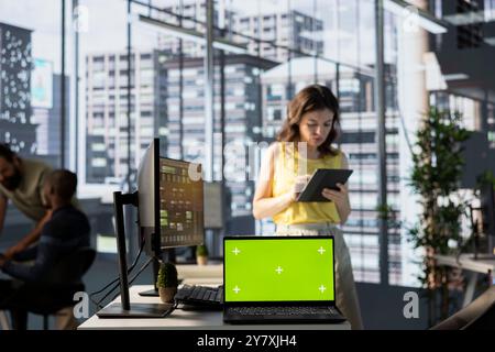Green screen laptop in front of admin doing software quality assurance, checking source code on tablet. Chroma key notebook in office with woman at work inspecting coding on device Stock Photo