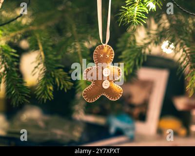 A handcrafted gingerbread-shaped ornament with white buttons hangs from a Christmas tree branch, illuminated by soft festive lighting. The background Stock Photo