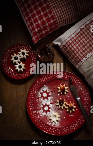 Christmas cookies (Spitzbuben) with jam, knife and on red plates, on wooden background Stock Photo
