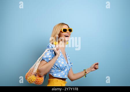 Fashionable young woman carrying mesh bag with oranges on shoulder against blue background Stock Photo