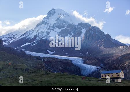 Georgian wonder: Mount Kazbek looms over alpine meadows, its icy crown contrasting with a sun-drenched refuge. Stock Photo