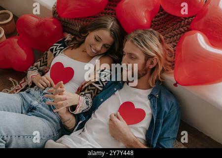 Top view of loving couple holding heart shape Valentines cards while leaning on bed surrounded with red balloons Stock Photo