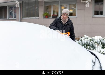 A mature man cleaning snow from car windshield outdoors on winter day. Concept of winter weather, car maintenance, and being prepared for snowy condit Stock Photo