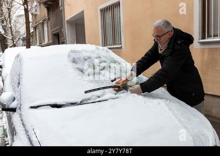 A mature man cleaning snow from car windshield outdoors on winter day. Concept of winter weather, car maintenance, and being prepared for snowy condit Stock Photo