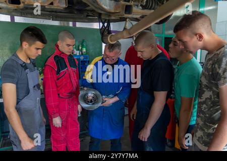 Mature experienced teacher standing underneath car on hydraulic ramp and teaching automotive trade to his students in the vocational school Stock Photo