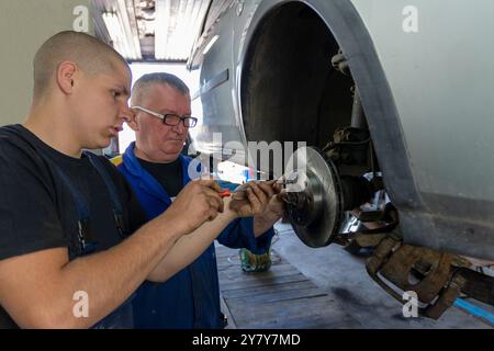 Mature experienced teacher standing underneath car on hydraulic ramp and teaching automotive trade to his students in the vocational school Stock Photo