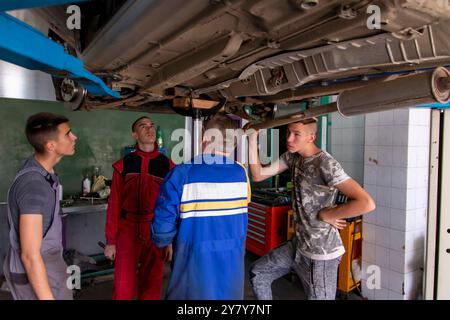 Mature experienced teacher standing underneath car on hydraulic ramp and teaching automotive trade to his students in the vocational school Stock Photo