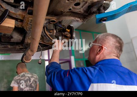 Mature experienced teacher standing underneath car on hydraulic ramp and repairing the car Stock Photo