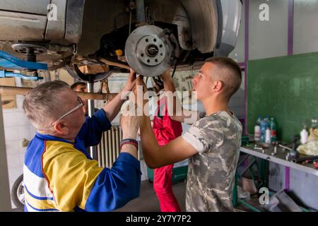Mature experienced teacher standing underneath car on hydraulic ramp and teaching automotive trade to his students in the vocational school Stock Photo