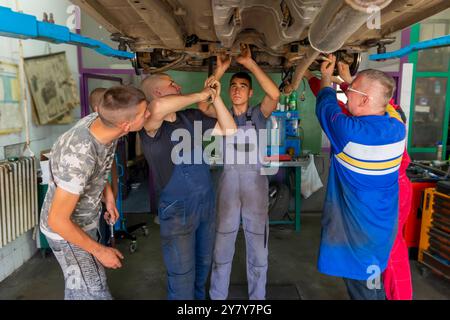 Mature experienced teacher standing underneath car on hydraulic ramp and teaching automotive trade to his students in the vocational school Stock Photo