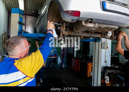 Mature experienced teacher standing underneath car on hydraulic ramp and teaching automotive trade to his students in the vocational school Stock Photo