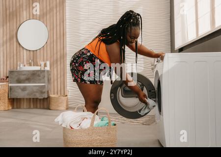 Beautiful curvy African woman loading clothes into washing machine while standing in bathroom Stock Photo