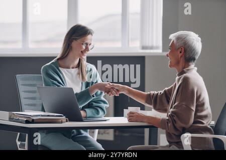 Happy senior woman shaking hands with financial advisor while sitting in office together Stock Photo