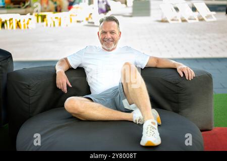 A middle-aged man lounges comfortably on a large outdoor sofa, dressed in a casual white t-shirt, grey shorts, and sneakers. Stock Photo