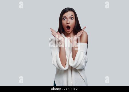 Oh my God! Shocked young woman keeping mouth open while standing against grey background Stock Photo