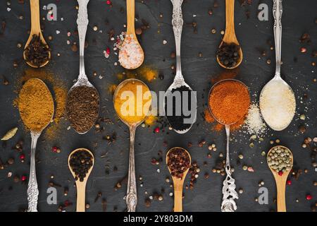 Many different exotic spices on tablespoons of metal and small wooden spoons arranged in a row in horizontal, landscape format on a large slate plate Stock Photo