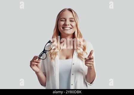 Attractive young woman pointing you and winking with smile while standing against grey background Stock Photo