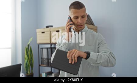Young man in office checking time on smartwatch while talking on phone, with office furniture and plant in background Stock Photo