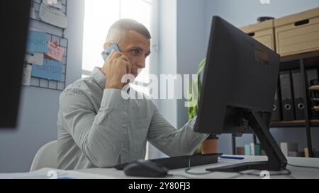 Young man working in office on computer while talking on phone, focused on monitor, indoors in modern workplace with documents and folders in backgrou Stock Photo