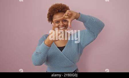 Smiling african woman framing face with fingers against a pink background, expressing creativity and joy. Stock Photo