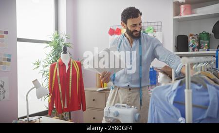 Bearded tailor man holding patterns in a bright atelier with mannequin and colorful fabrics. Stock Photo