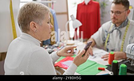 Two tailors collaborate in a design room, with a woman holding a phone and fabric samples on the table. Stock Photo