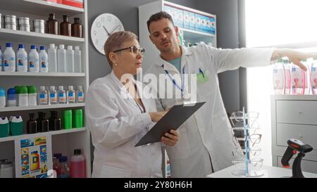 Woman and man pharmacists consulting clipboard in pharmacy with shelves filled with medicines and products around them. Stock Photo